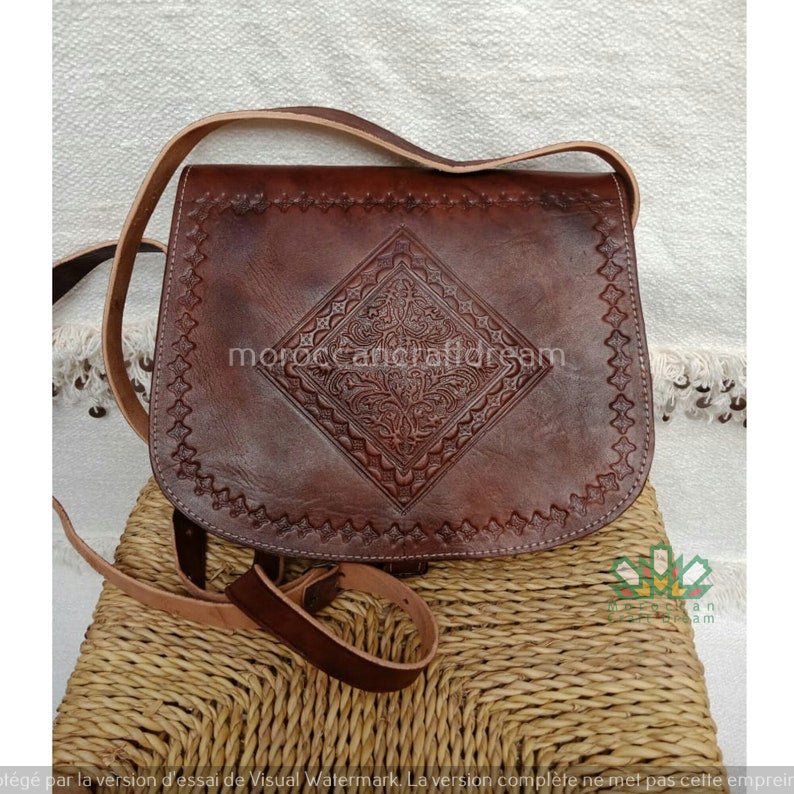 A handmade dark tan leather bag with intricate embossing, displayed on a woven mat.