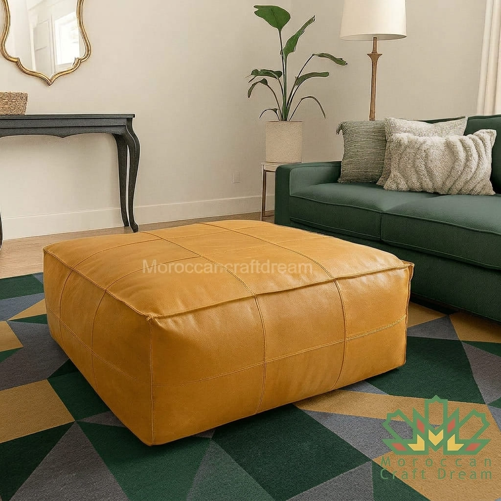 A square mustard yellow leather pouf ottoman with stitched panel seams, placed on a geometric green and beige rug in a styled living room next to a green sofa.