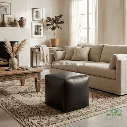A styled living room scene featuring a beige fabric sofa, a black leather cube ottoman, a carved wooden coffee table, and a patterned area rug in a warm, neutral-toned interior.