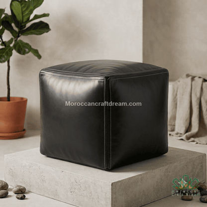 A black cube-shaped leather pouf ottoman displayed on a concrete pedestal in a styled room setting with a potted plant and draped fabric in the background.