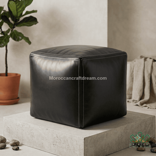 A black cube-shaped leather pouf ottoman displayed on a concrete pedestal in a styled room setting with a potted plant and draped fabric in the background.