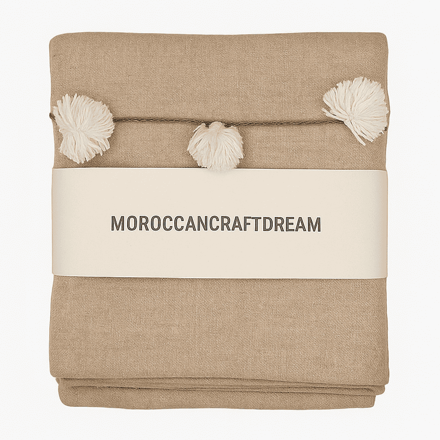 Beige blanket with pom-poms and 'MOROCCANCRAFTDREAM' label on a white background