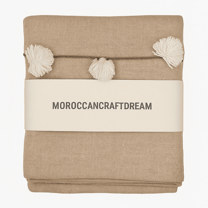 Beige blanket with pom-poms and 'MOROCCANCRAFTDREAM' label on a white background