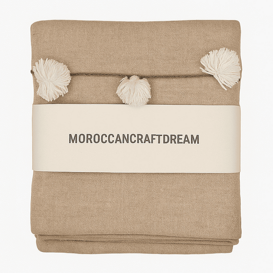 Beige blanket with pom-poms and 'MOROCCANCRAFTDREAM' label on a white background