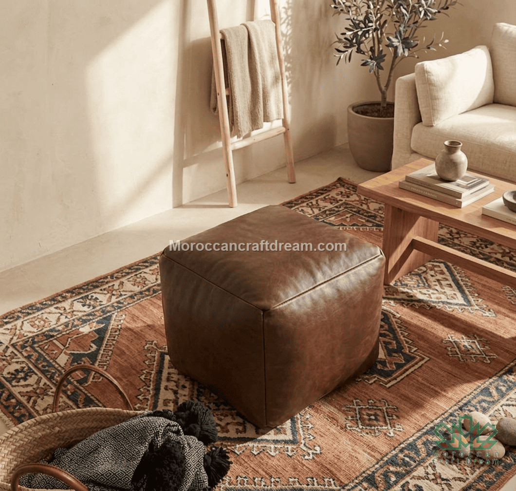A square brown leather ottoman placed on a patterned area rug in a styled living room setting with a wooden coffee table, sofa, and ladder shelf in the background.