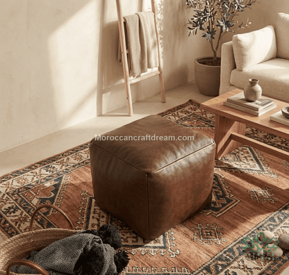 A square brown leather ottoman placed on a patterned area rug in a styled living room setting with a wooden coffee table, sofa, and ladder shelf in the background.