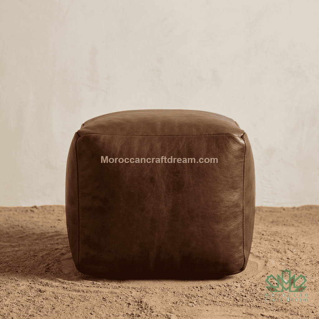A square chocolate leather pouf ottoman with a smooth texture, set against a neutral beige textured wall and sandy floor. A watermark 'Moroccancraftdream.