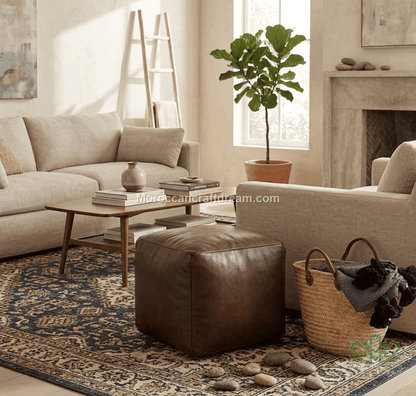 A styled living room scene featuring a brown leather ottoman in the foreground, a beige sofa and armchair, a patterned area rug, a wooden coffee table, a potted fiddle-leaf fig, and a woven basket on a stone floor near a fireplace.