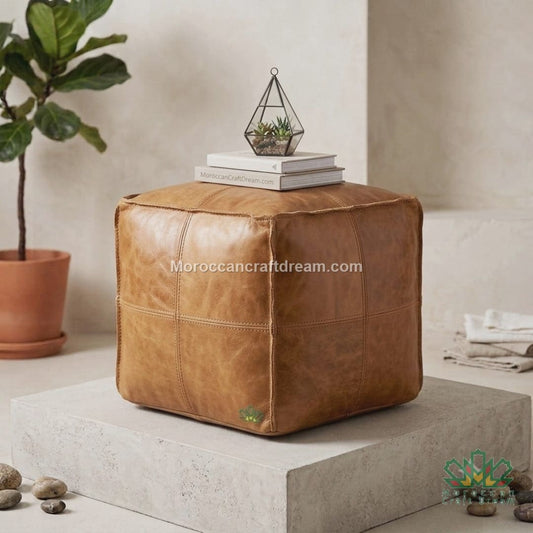 A square darky light caramel leather pouf ottoman with stitched paneling, styled on a concrete pedestal with books, a small plant in a glass terrarium, and a potted fiddle-leaf fig in a neutral room setting.