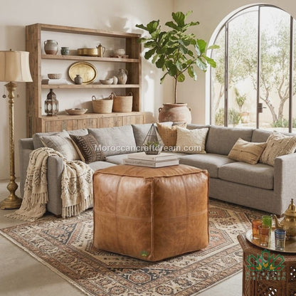 A styled living room featuring a central light caramel leather cube pouf ottoman on a patterned rug, set in front of a gray sectional sofa with a wooden shelving unit and a large arched window in the background.