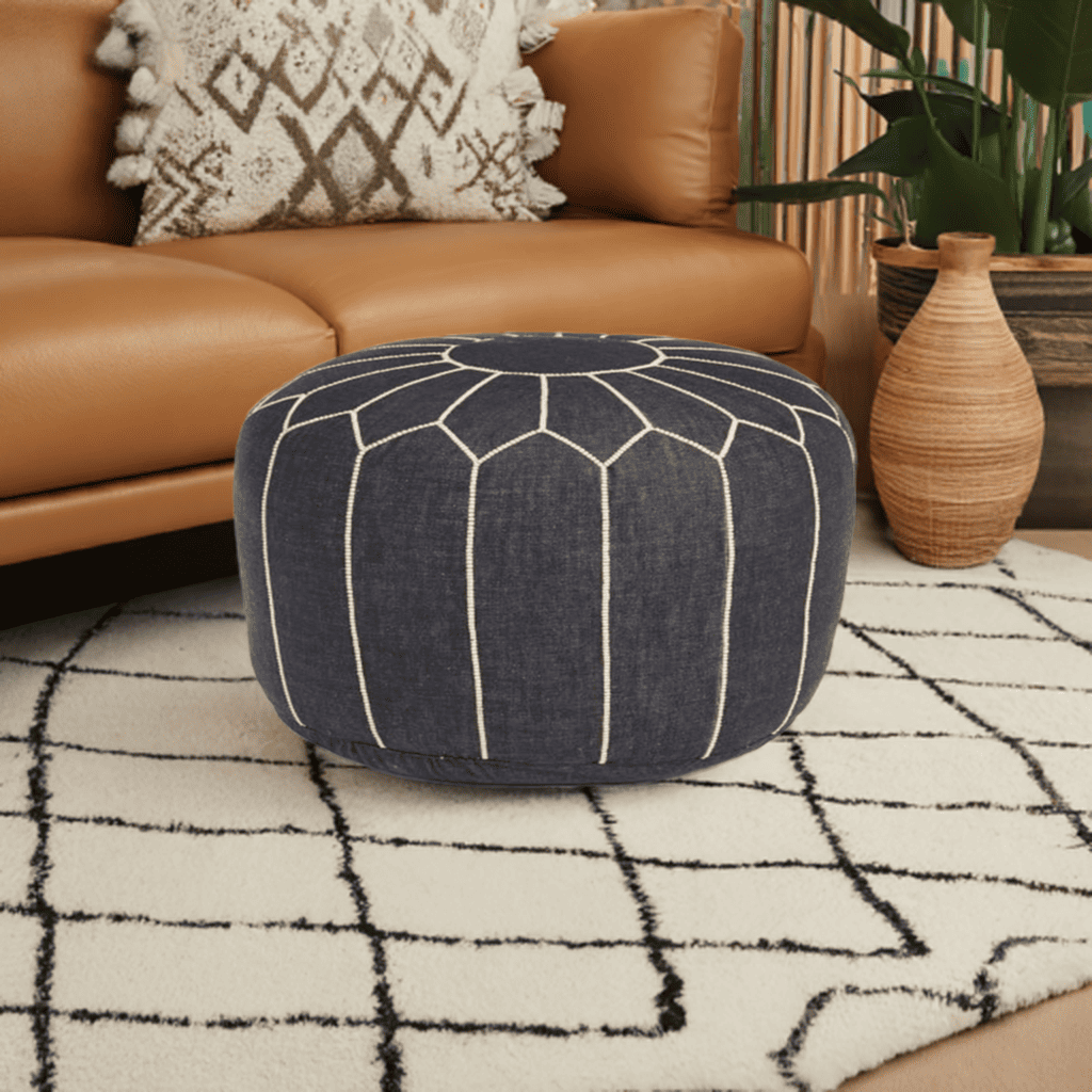 A round navy blue denim pouf with white hand-stitching patterns, placed on a rug outdoors, with a chair and potted plant in the background.