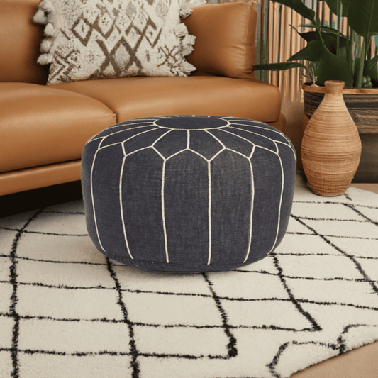 A round navy blue denim pouf with white hand-stitching patterns, placed on a rug outdoors, with a chair and potted plant in the background.