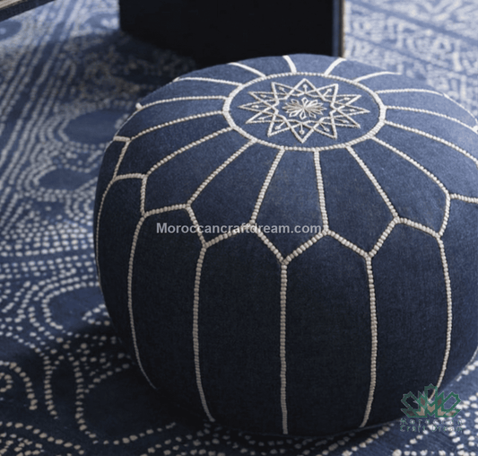 A round denim Moroccan pouf ottoman in dark navy blue fabric with white embroidered geometric patterns, shown on a matching patterned rug.