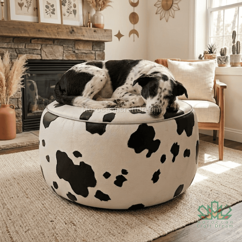 A black and white cow-print round ottoman with a thick cushion, shown in a cozy living room with a black and white dog sleeping on top. The background features a stone fireplace, a wooden chair, and a neutral rug.