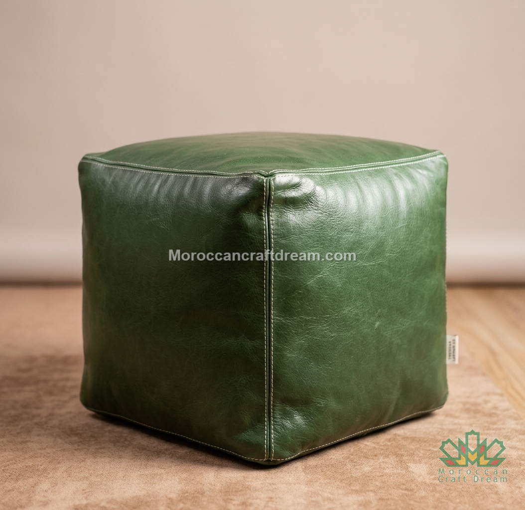 A square green leather pouf ottoman with stitched panel seams, shown on a wooden floor against a neutral beige wall. A watermark 'Moroccancraftdream.