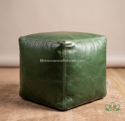 A square green leather pouf ottoman with stitched panel seams, shown on a wooden floor against a neutral beige wall. A watermark 'Moroccancraftdream.