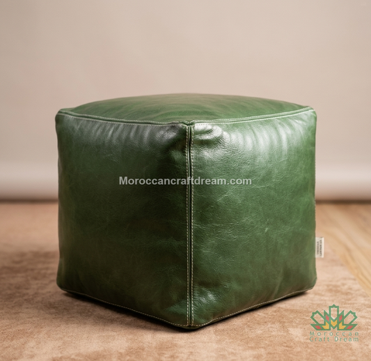 A square green leather pouf ottoman with stitched panel seams, shown on a wooden floor against a neutral beige wall. A watermark 'Moroccancraftdream.