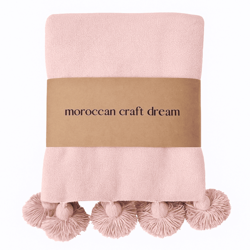 Pink blanket with pom-pom trim and 'moroccan craft dream' label on a white background