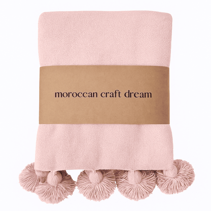 Pink blanket with pom-pom trim and 'moroccan craft dream' label on a white background