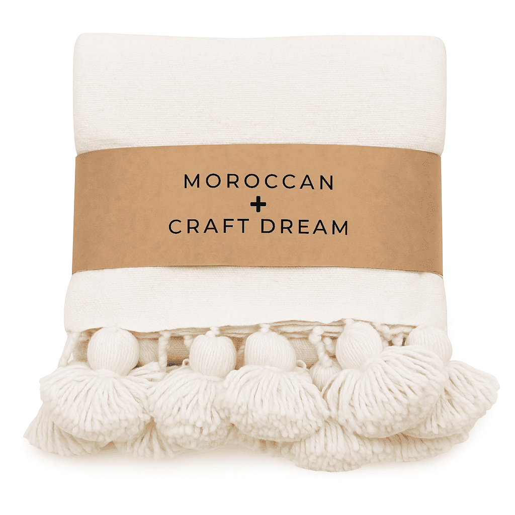 White blanket with pom-poms and a brown label reading 'Moroccan Craft Dream' on a white background.