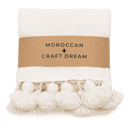 White blanket with pom-poms and a brown label reading 'Moroccan Craft Dream' on a white background.