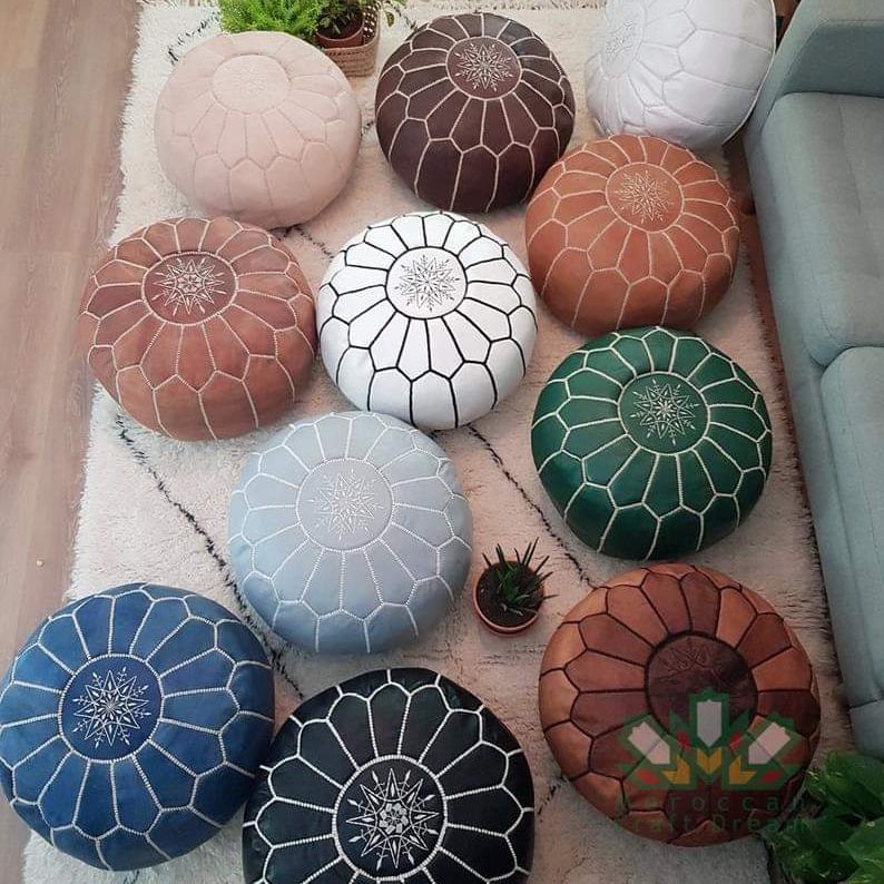 A set of round Moroccan-style pouf ottomans in multiple colors including blue, black, pink, brown, white, orange, green, and light blue, arranged on a white rug next to a sofa.