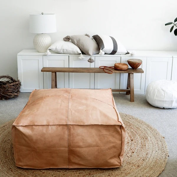 A large square Moroccan leather coffee table ottoman in a neutral color, placed on a round area rug next to a white bench with cushions, in a room with a white cabinet and decorative items.