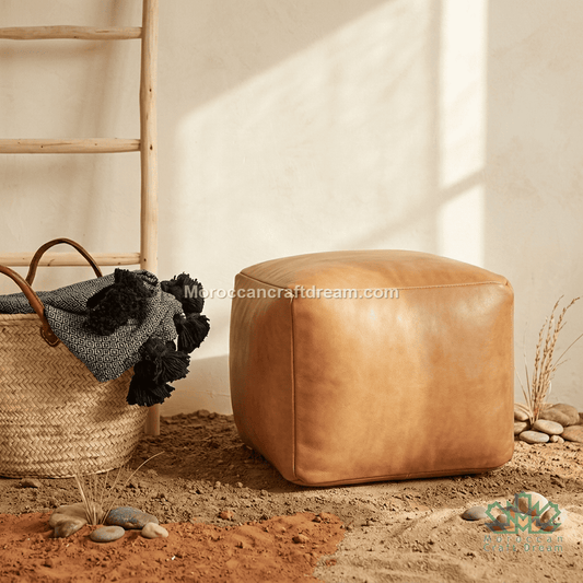 A square tan leather pouf ottoman sits on a sandy floor next to a woven basket with a dark knit throw, against a warm off-white wall with soft window shadows.