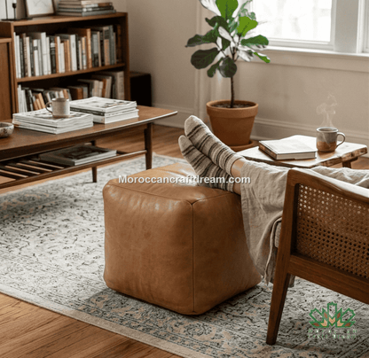 A tan leather cube ottoman in a cozy, sunlit living room setting next to a wooden chair with a woven cane backrest. A beige throw blanket is draped over the ottoman, and a striped pillow rests on top.