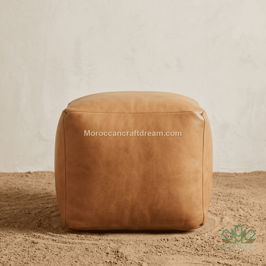 A square tan leather pouf ottoman with a smooth, slightly distressed finish, set on a sandy surface against a textured off-white wall. A watermark 'Moroccancraftdream.