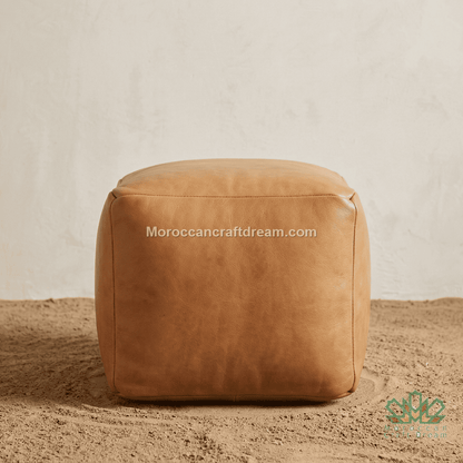 A square tan leather pouf ottoman with a smooth, slightly distressed finish, set on a sandy surface against a textured off-white wall. A watermark 'Moroccancraftdream.