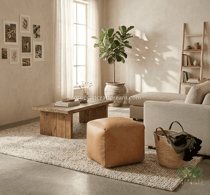 A styled living room scene with a rustic wooden coffee table, tan leather ottoman, beige sofa, woven rug, potted fiddle-leaf fig, basket, and framed wall art in a warm neutral interior.