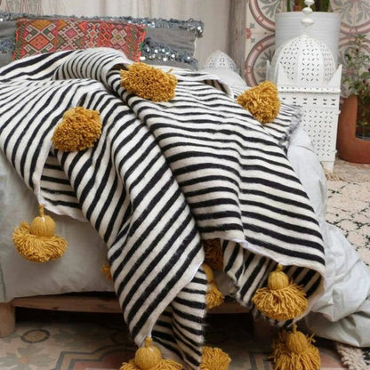 A black and white striped Moroccan throw blanket with yellow pom pom details, draped over a sofa in a room with bohemian decor.