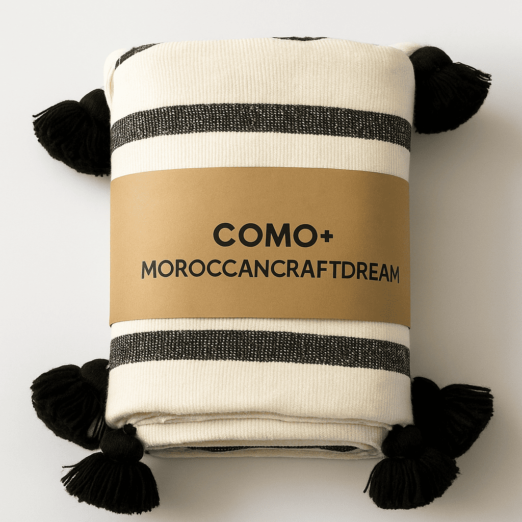 Folded blanket with black and white stripes and pom-poms, featuring 'COMO+ MOROCCAN CRAFT DREAM' label on a light gray background.