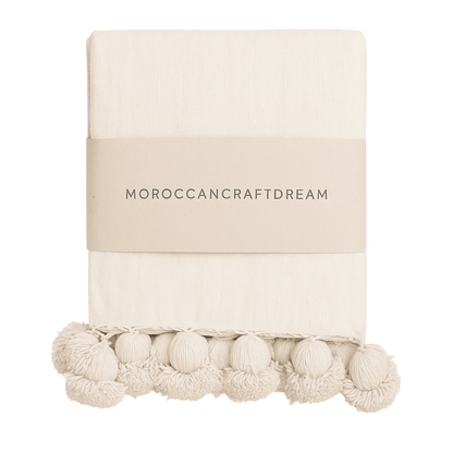 Beige throw blanket with pom-pom trim on a white background, featuring 'Moroccancraftdream' branding.