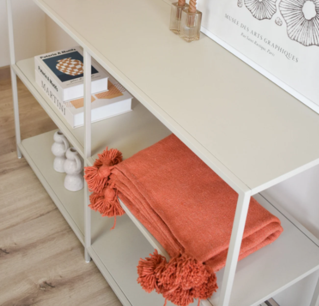 White shelf with books, a bottle, and an orange blanket with pom-poms on a wooden floor.