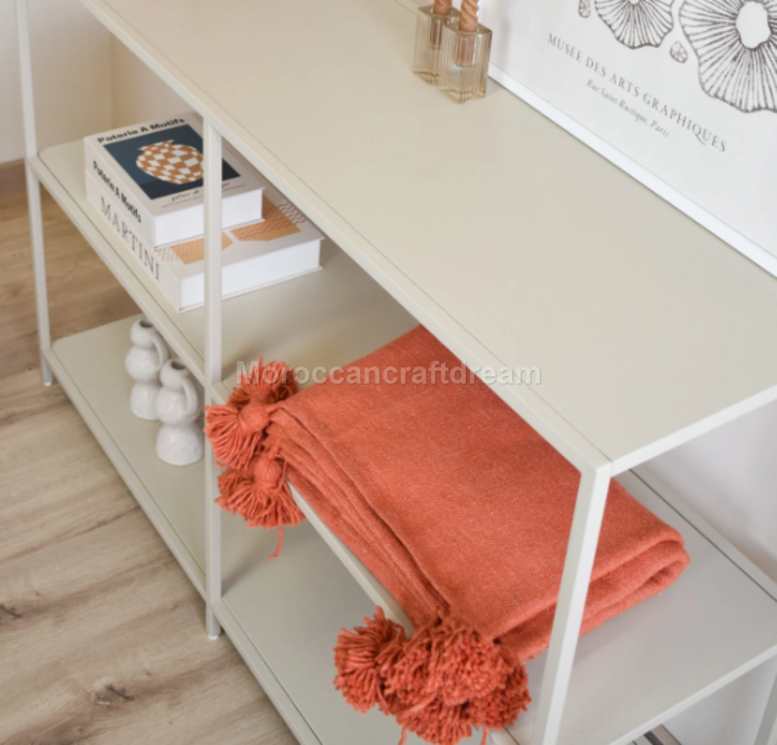 White shelf with books, a framed picture, and an orange throw blanket on a wooden floor.