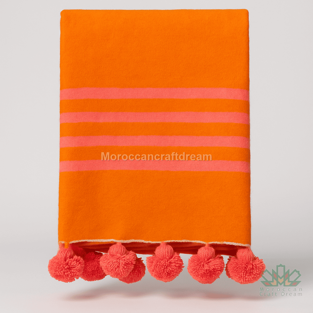Orange handmade blanket with pink stripes and pom-poms on a light gray background