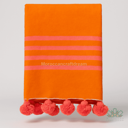 Orange handmade blanket with pink stripes and pom-poms on a light gray background