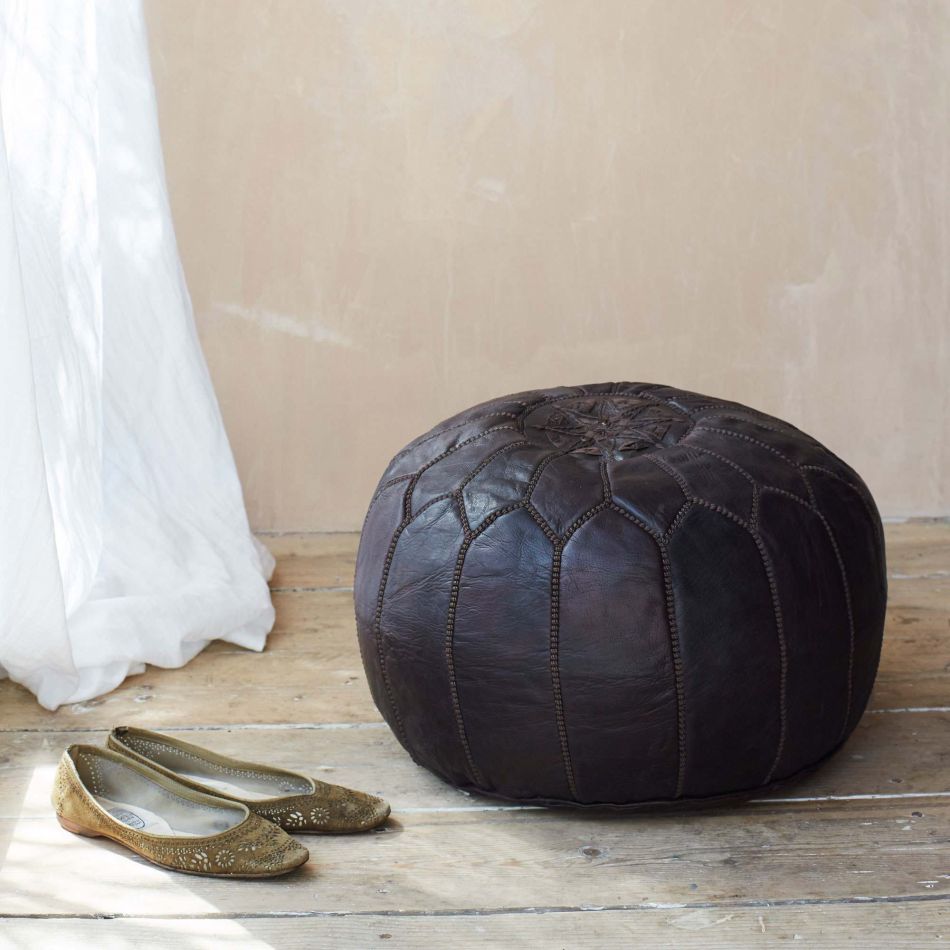 A handmade black round leather Moroccan pouf placed on a wooden floor against a wall, next to a pair of gold-colored shoes.