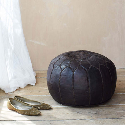 A handmade black round leather Moroccan pouf placed on a wooden floor against a wall, next to a pair of gold-colored shoes.