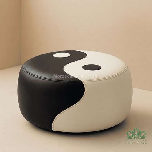 A round pouf ottoman upholstered in black and off-white leather with a yin-yang design, set against a neutral beige background.