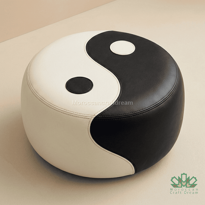 A round pouf ottoman with a yin-yang design, featuring black and off-white faux leather panels, photographed on a neutral beige background.
